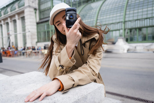 young woman in trench coat and baseball cap taking photo on vintage camera.