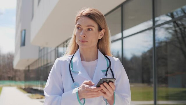 Female doctor physician with a stethoscope standing near clinic outdoors holding mobile phone texting message scrolling screen of smartphone sends online prescription to the patient during a pandemic.
