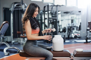 An attractive sportswoman is making a protein shake while sitting on a bench in a gym.