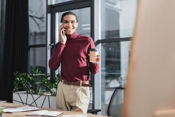 Smiling african american businessman talking on smartphone and holding coffee to go near notebook in office.