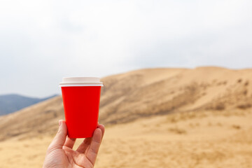 A red cup of coffee or a hot drink, covered with a lid in your hand against the background of a desert or a sandy seashore.