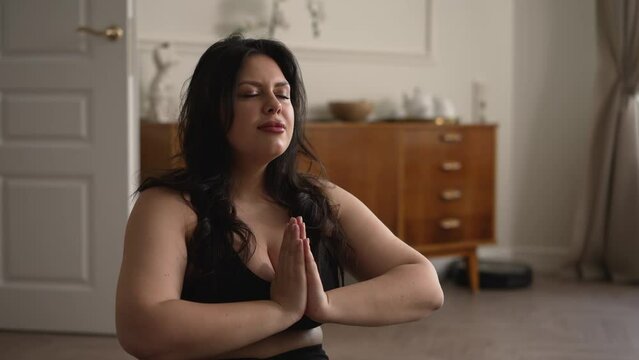 Young overweight woman meditating and doing yoga on floor in apartment room spbd. 4k Close view Caucasian beautiful brunette woman does asana with closed eyes and controls breath, meditates with smile