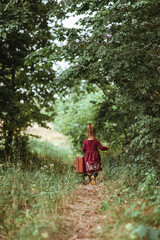 Rear view of a pretty little girl in burgundy dress holding her old suitcase and walking in the forest at summer time, retro style