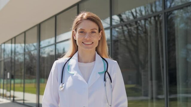 Close up portrait of caucasian female healthcare worker doctor standing outside hospital turning head smiling. Confident face of surgeon wearing scrubs and stethoscope posing on camera. Real emotion.
