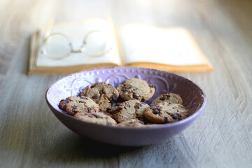 Bowl of chocolate chip cookies, open book and reading glasses on the table. Selective focus.