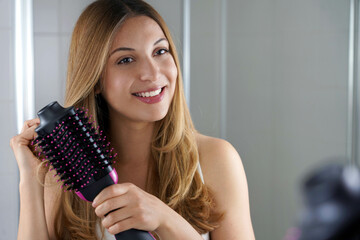 Pleased girl holds round brush hair dryer to style hair in her bathroom at home. Young woman using...