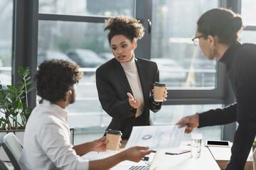 African american businesswoman holding coffee while talking to multiethnic colleagues with paper in office.