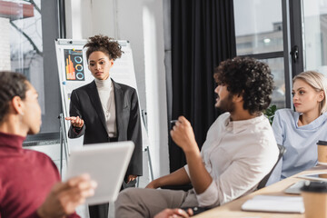 African american businesswoman pointing at colleague near multicultural business people and flip chart in office.