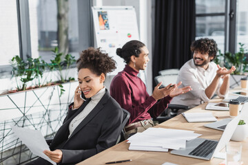 Positive african american businesswoman talking on smartphone and holding paper near blurred interracial colleagues in office.