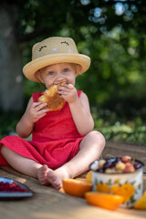  A little girl in a straw hat and a red dress with bare feet eats a croissant
