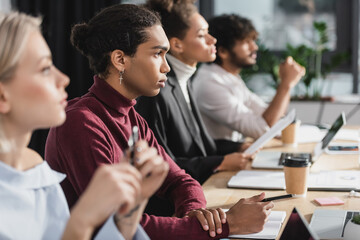 Side view of african american businessman looking away near blurred multiethnic colleagues at table in office.