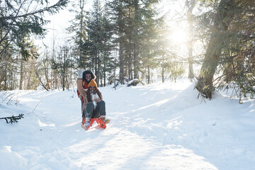 Fototapeta na wymiar Long shot of playful young Black man pushing his joyful girlfriend on sledge in forest on sunny winter day