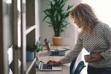 Adult businesswoman working on laptop sending email. Standing woman work on computer writing while drink a coffee. Home job occupation for digital online remote worker. Smart working concept lifestyle