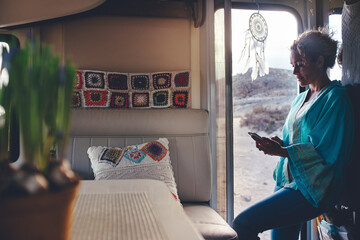 Adult woman smile and use mobile phone to send message standing on the door of her alternative house camper van. Tourist enjoying travel with caravan and using roaming connection - obrazy, fototapety, plakaty