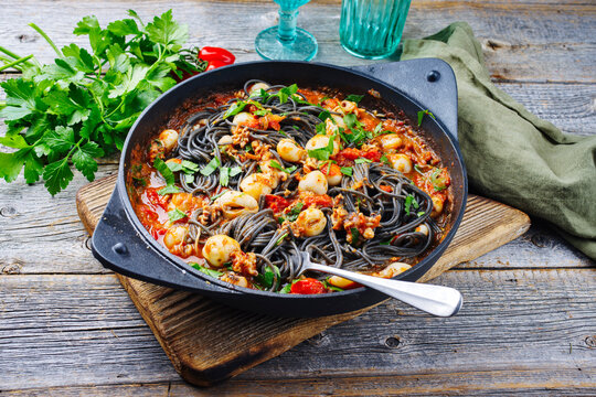 Traditional Italian spaghetti al nero di seppia with baby squid ink in tomato sauce served as close-up in a cast iron pan on a wooden board