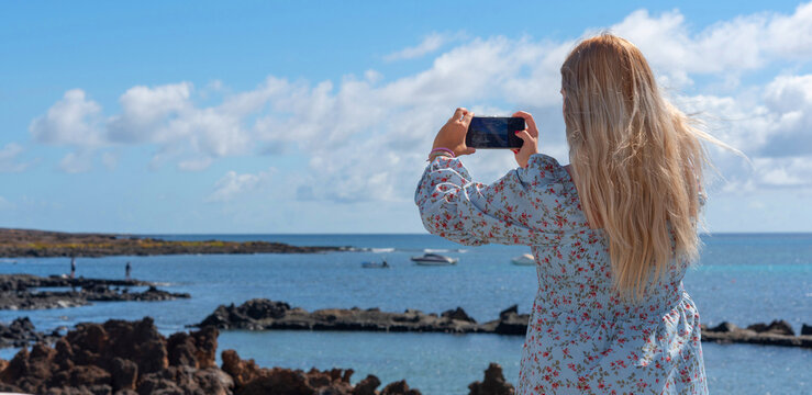 Una mujer joven rubia de espaldas sacando una foto con el teléfono móvil en la costa de Lanzarote en las Islas Canarias. Turismo veraniego. 