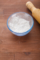 Confectionery flour in a bowl on a wooden table next to a rolling pin.