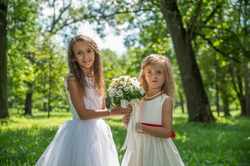 Two little girls sisters in white dresses in the park among the summer greenery in the backlight of the sun.