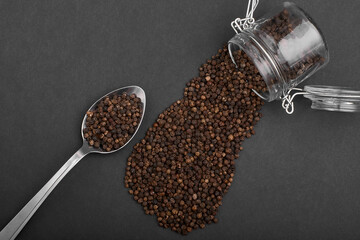 granules of small allspice in a spoon and scattered from a jar on a black background
