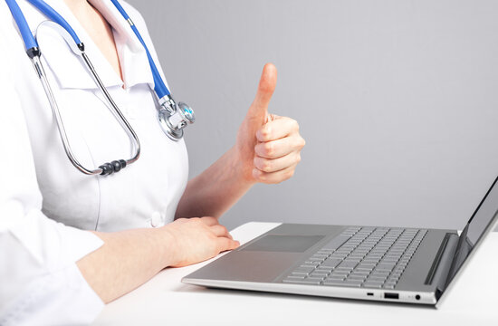 Doctor giving online consultation to patient and showing thumb up gesture. Telemedicine, telehealth concept. Woman with stethoscope sitting at table with laptop and using video chat. photo