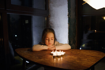 a little girl with her eyes closed is sitting at a table in front of burning candles. The child...