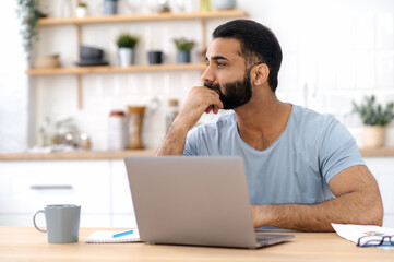 Fototapeta na wymiar Thoughtful handsome indian or arabian man in casual t-shirt, freelancer, designer, working from home, sitting in the kitchen with a laptop, looking away, thinking about a new project, strategy