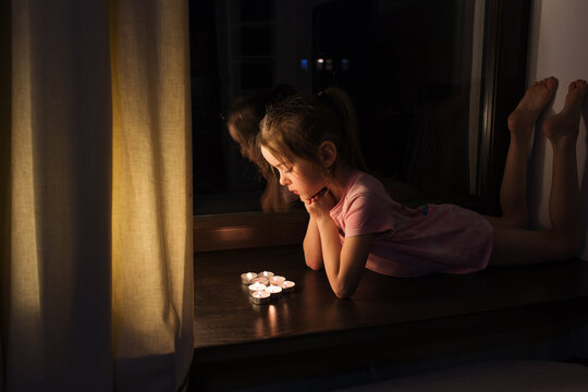 a little girl in a T-shirt lies on the windowsill near the window and looks at the burning candles