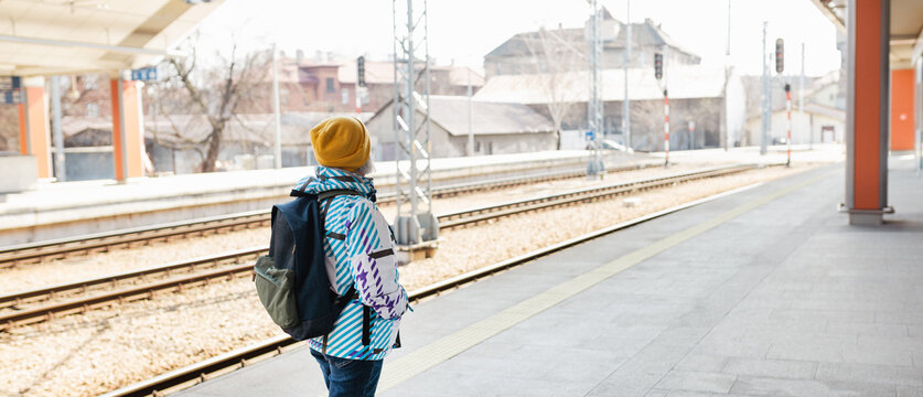 Old happy woman with backpack in yellow hat waiting train on station platform on urban background. Railroad transport concept