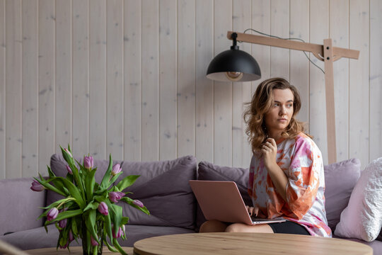 Portrait Of Woman Working On Modern Laptop Sitting On Soft Violet Sofa In Casual Clothes Near Table With Vase Of Fading Tulips. Living Room In Elegant Design With Aesthetic Decor, Free Copy Space