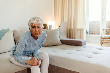Fototapeta premium A woman suffering from pain in the knee, closeup. Old age, health problem and people concept - mature woman suffering from pain in leg at home. Photo of senior woman holding the knee with the pain.