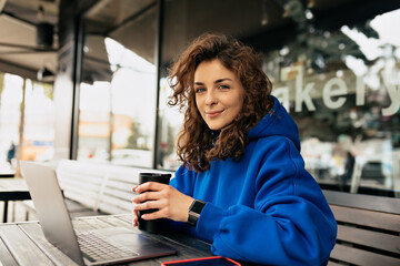 Close-up portrait of inspired pretty lady with curly hairstyle is working on laptop with coffee in the morning. Outdoor photo of smiling shy girl is working remote in the city in morning