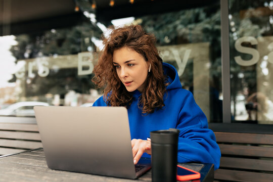focused cute girl with curls wearing oversize blue pullover is working on laptop while sitting on outdoor cafe with coffee