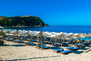 Landscape of beautiful clean sand and pebble beach with beach umbrellas and sun loungers on mountains background. Himare. Albania.