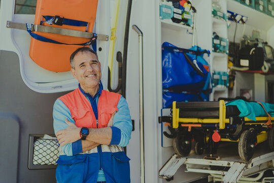 corporate portrait of a smiling middle-aged doctor with his arms crossed, standing next to an ambulance with open doors.