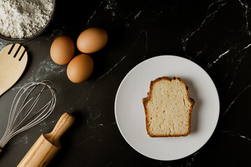 Aerial photo of a black marble work table with eggs, a whisk, a rolling pin, flour and a slice of sponge cake on a plate.