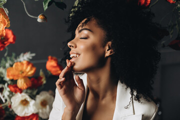 Studio shot of overjoyed dark skinned female model laughs happily and poses near flowers in white...