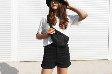 Woman, wearing white t-shirt, black shorts, fanny pack or waist pack and bucket hat, standing outdoor near white wall. Details of stylish trendy basic minimalistic casual outfit. Street fashion. 