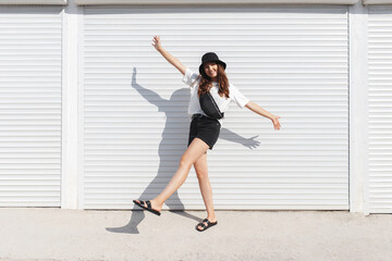Woman, wearing white t-shirt, black shorts, fanny pack or waist pack, bucket hat and flat sandals, standing outdoor near white wall. Stylish trendy basic minimalistic casual outfit. Street fashion.