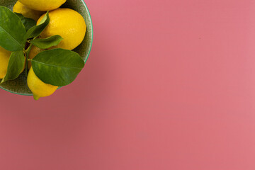 Overhead shot of a bowl full of fresh organic lemons with their green leaves on a pink table