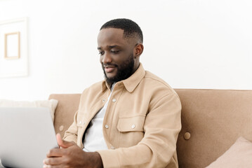 Image of happy afro american man working on laptop, watching social media news or doing online shopping while sitting in living room. Young smiling black freelancer working from home