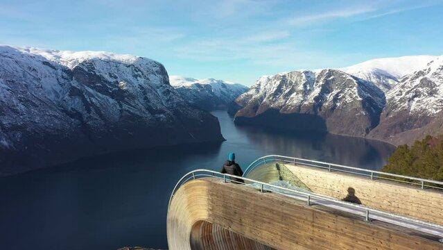 Flying slowly above man relaxing on edge of spectacular Stegastein viewpoint - Aurlandsfjorden and sognefjord in background - Norway winter aerial