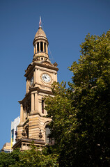 Fototapeta na wymiar Clock tower at Town Hall, Sydney, Australia. Set against a deep blue sky and framed with green trees on the left. Time 10.45am. Hand wound clock with 4 glass clock faces. 