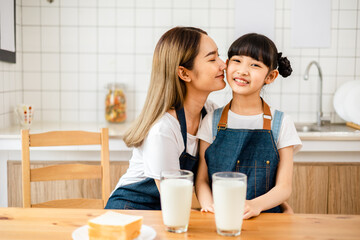 Asian teen girl drinking milk with her mom and eating bread, sitting at kitchen table.Loving mom kissing her little daughter in the kitchen, cheerful family .