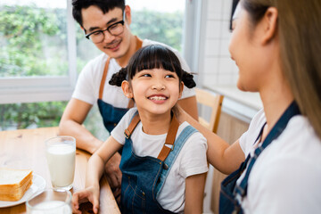 Asian  family enjoying breakfast at cozy kitchen, little girl daughter sitting on table, drinking milk with smiling father and mother in morning. Happy family in kitchen.