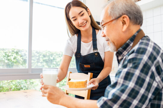 Asian Lovely Family, Young Daughter Prepare Breakfast For Older Father. Attractive Female Wear Apron Bake Bread Serve With Milk To Senior Elderly Dad Sitting On Eating Table In Kitchen At House.