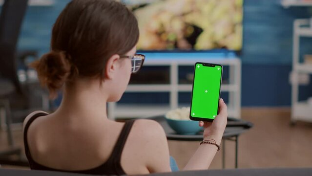Closeup of young woman holding vertical smartphone with green screen in online conference or group video call in home living room. Static shot of girl using touchscreen mobile phone with chroma key.