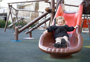 Cute adorable pretty caucasian blonde baby girl sliding,having fun.Happy, warm clothing child,kid,toddler, infant of 1-2 year old in playground .Lifestyle outdoors, copy space for text
