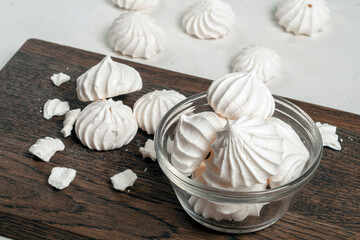 French Meringue dessert lies in a glass bowl, on a dark wooden board. there are whole and broken meringues lying nearby. Meringue is made from a mixture of beaten egg whites with sugar.