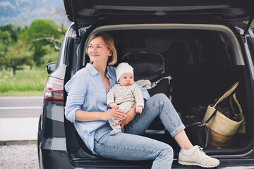 Mother with baby sitting in open car trunk outdoors. Road trip with baby. Travel in Europe. Young...