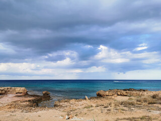 The sandy coast of the Mediterranean Sea, turning into stones from a long-hardened lava, azure water against a dramatic sky.